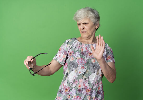 老人恼怒的老妇人举着眼镜举起手隔离在绿墙上举行手眼镜