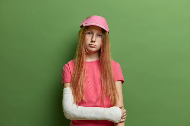 情绪小女孩手臂骨折 面部表情伤感 戴帽子 穿休闲t恤 骨头有问题 皮肤有雀斑 隔离在绿色墙壁上儿童和受伤概念疾病事故疼痛