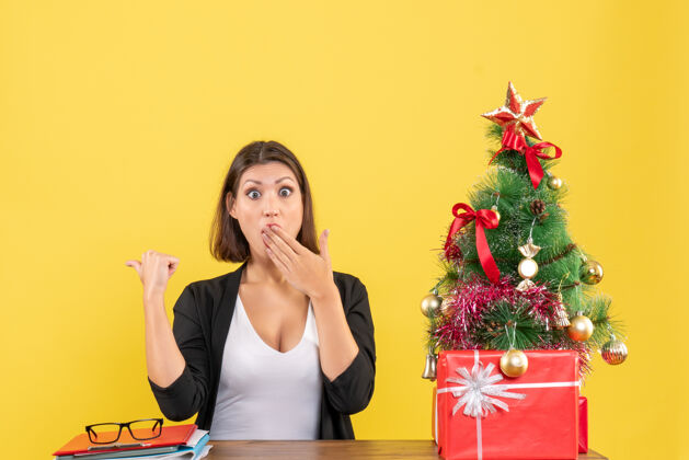 圣诞一位穿着西装的年轻女士惊讶地指着黄色办公室装饰好的圣诞树可爱封面女郎漂亮