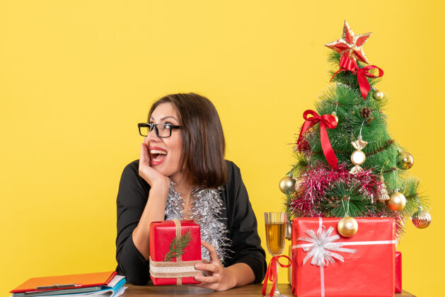 成人一位穿着西装 戴着眼镜展示礼物 坐在办公室里一棵圣诞树旁的商务女士大吃一惊导演桌子时尚