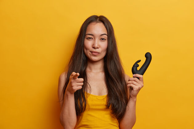 亲密自信的年轻女子指着食指 手持振动器刺激阴蒂闪烁的振动 有个人假阴茎 隔离在黄色墙壁上女性性玩具兔子橡皮内部
