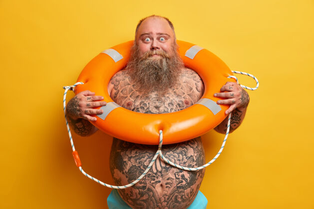 帅气夏季休息和安全游泳的概念震惊的胡子粗壮的男人赤裸着站着 有纹身的身体和大肚子 摆着充气的救生圈 等待假期 隔离在黄色的墙壁上人肥胖赤膊