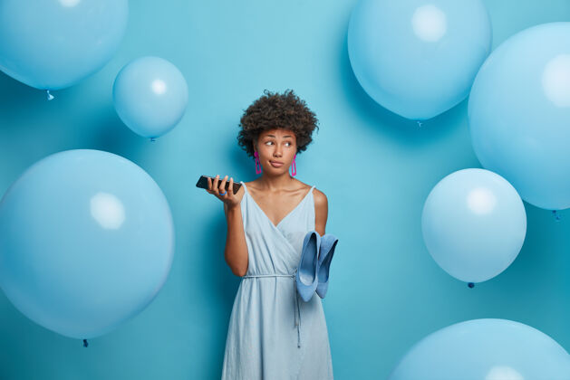 优雅可爱迷人体贴的年轻美国黑人女子准备约会 手里拿着智能手机 等待男友的电话 穿着蓝色的裙子和鞋子 站在室内靠近气球充气服装衣柜