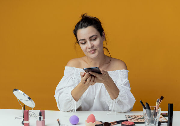 坐着高兴的漂亮女孩坐在桌子旁 拿着化妆工具 看着隔离在橙色墙上的手机请工具漂亮