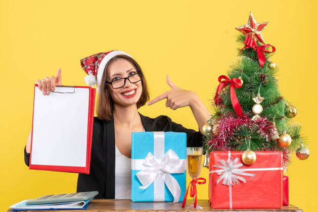 圣诞老人惊讶迷人的女士穿着西装戴着圣诞老人的帽子看着文件指着自己在办公室里的黄色隔离笔记本电脑人快乐