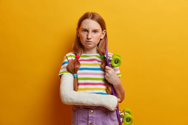恼怒恼火的小女孩 姜黄色头发和雀斑 笑脸和不满意的表情 与滑板姿势 不能继续驾驶 因为手臂创伤儿童 保健 危险的运动恼怒绷带室内