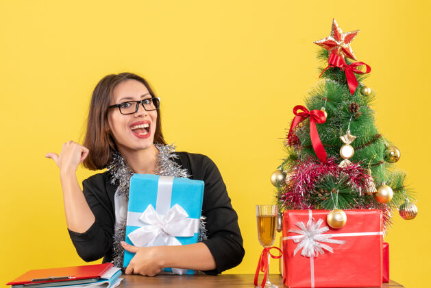 抱着一位身着西装 戴着眼镜 面带微笑的商务女士 手拿礼物 指着身后 坐在办公室的一张桌子旁 桌上放着一棵圣诞树时尚礼物微笑
