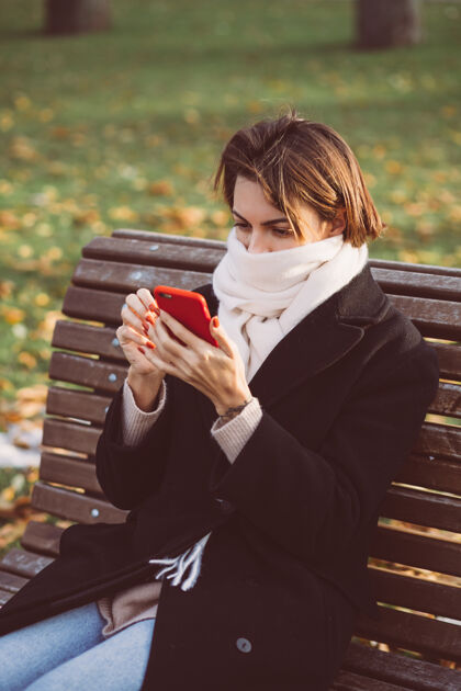 冷穿着黑色冬衣 戴着围巾 坐在公园长凳上 拿着手机的白种女人的户外画像移动围巾外套