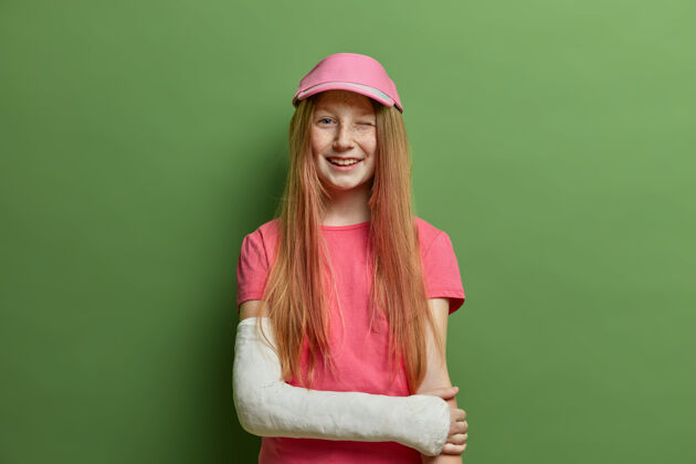 受伤儿童和保健概念快乐的红发女孩摆姿势 手臂骨折 在路上摔倒或意外受伤 穿着夏季t恤和帽子 眨眼 忘记外伤伤害保险白种人