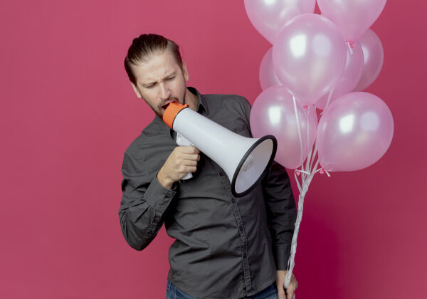大声恼羞成怒的帅哥拿着氦气球站在粉红色的墙上对着隔离的扩音器大喊气球喇叭站着