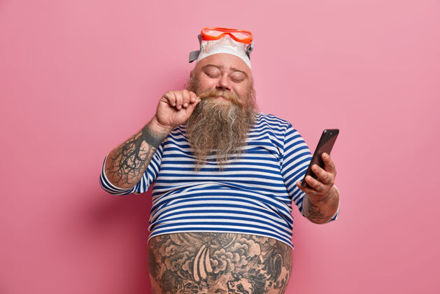 手机高兴的超重男子闭着眼睛站着摸胡子拿着智能手机看新闻 戴着泳镜和条纹水手衬衫享受良好的休息游泳运动员准备潜水颜色超重胡子
