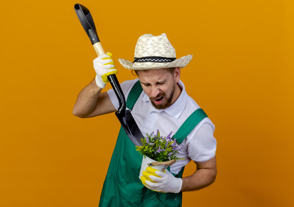 年轻人恼怒的年轻英俊的斯拉夫园丁穿着制服戴着帽子和园艺手套手持铁锹和花盆在花盆里铲花帅哥花园花