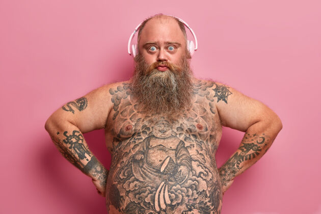 腹部科技和生活理念严肃自信的胖男人戴着耳机 听音乐 有着纹身的身体 肥胖的肚子 浓密的胡须 对着粉色的墙壁摆姿势 找到了很棒的播放列表耳机男人脂肪