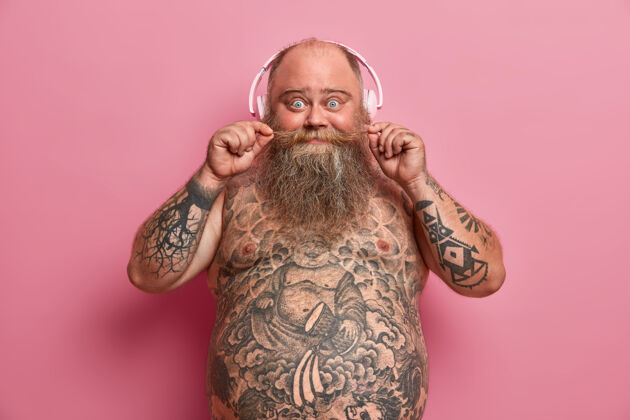 胡须惊讶有趣的胖胖男人留着胡子 赤膊上阵站在粉红色的墙上 有着肥胖的肚子 肚子和胳膊上都有纹身 喜欢的音乐让他发冷 戴着耳机 享受新的音乐过量胃大