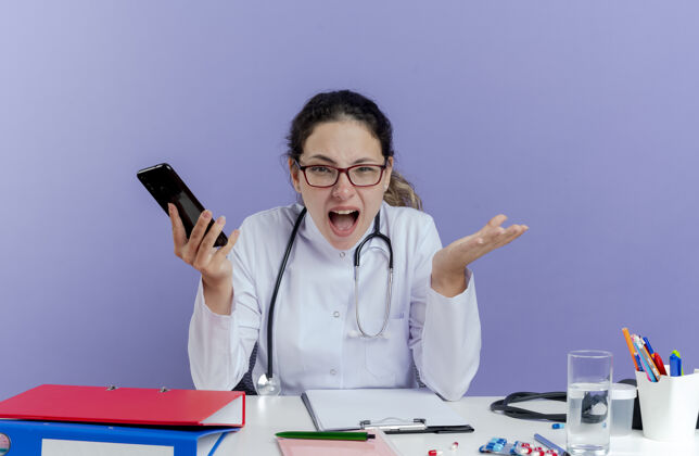 工具愤怒的年轻女医生穿着医用长袍 手持听诊器坐在办公桌旁 手持医疗工具 手持手机 显示空手孤立听诊器人展示