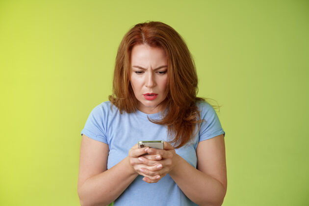 沮丧困惑不确定的红发中年妇女学习如何使用社交媒体尝试理解表情表情看起来紧张专注的智能手机显示阅读重要新闻信息站在绿色的墙红发紧张女人