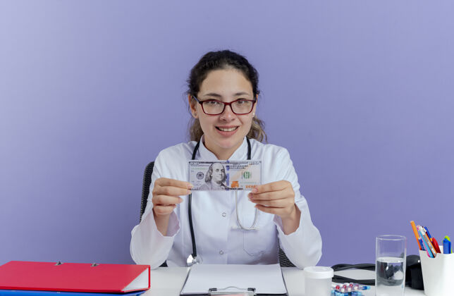 工具面带微笑的年轻女医生穿着医用长袍和听诊器坐在办公桌旁 手里拿着医疗工具 手里拿着钱 看上去与世隔绝听诊器桌子紫色