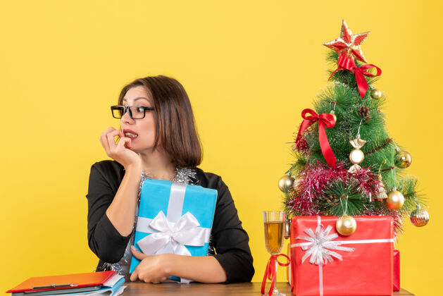 抱一位情绪激动的商务女士 穿着西装 戴着眼镜 手里拿着礼物 坐在一张桌子旁 桌子上放着一棵圣诞树坐树情绪