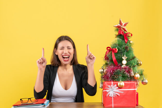 向上在黄色的办公室里 一个穿着西装的满意的年轻女人指着装饰过的圣诞树人欢呼年轻女子