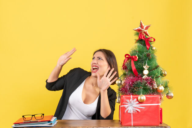 坐着情绪激动的年轻女子坐在黄色办公室装饰圣诞树旁的桌子旁看着什么东西快乐树人