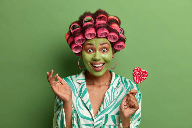 肖像快乐正面的非洲裔美国女人的横拍看起来很高兴 咯咯笑着接受美容治疗 敷上美容面膜 拿着美味的棒棒糖 戴着卷发器 隔离在绿色上粘土身体情绪