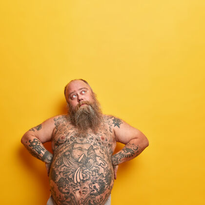 空胖胖体贴的男人双手叉腰 大裸着刺青的肚子 浓密的胡须 愁眉苦脸的向上看 表情严肃 想着如何减肥 孤身在黄色的墙上脂肪表情头发