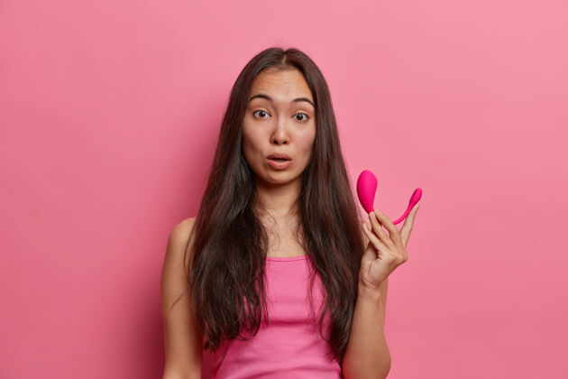 性惊讶的黑发女性手持现代远程应用程序控制的智能振动器 逛性用品店 购买必要的配件来满足自己 穿着随意 对着粉色的墙壁摆姿势印象情色表情