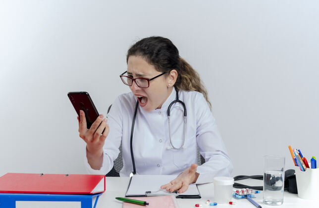 愤怒愤怒的年轻女医生穿着医用长袍 戴着听诊器和眼镜坐在办公桌前拿着医疗工具 孤立地看着手机穿长袍眼镜