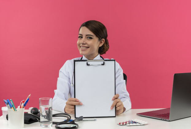 年轻快乐的年轻女医生穿着医用长袍和听诊器坐在办公桌旁 带着医疗工具和笔记本电脑剪贴板听诊器人
