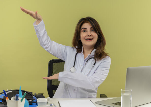 医疗令人印象深刻的中年女医生穿着医用长袍和听诊器坐在办公桌前 医疗工具剪贴板和笔记本电脑显示大小隔离医生尺寸个人