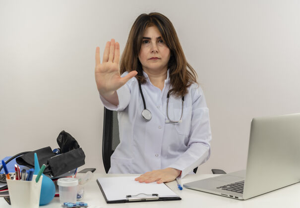 成人严格的中年女医生穿着医用长袍和听诊器坐在办公桌前用医用工具剪贴板和笔记本电脑做停止手势坐笔记本电脑严格