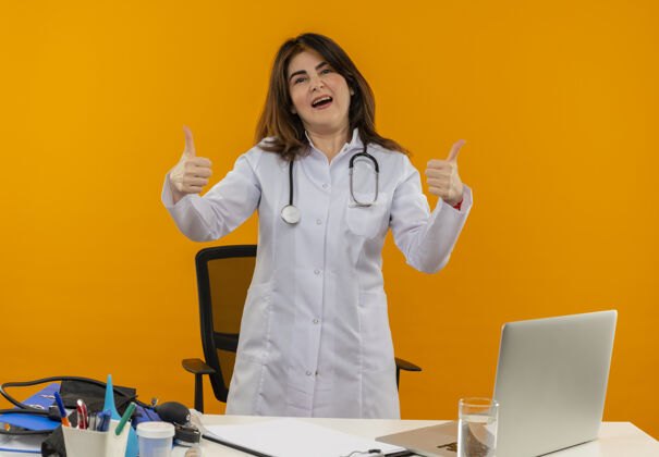 向上自信的中年女医生穿着医用长袍 听诊器站在桌子后面 带着医疗工具剪贴板和笔记本电脑 竖起大拇指女士长袍工具