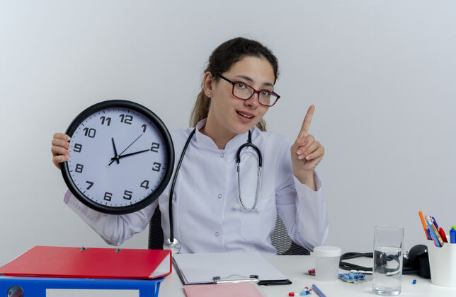 穿着年轻的女医生穿着医用长袍 戴着听诊器 戴着眼镜 坐在办公桌旁 手里拿着医疗工具 手握时钟 手指被隔离工具提高医生
