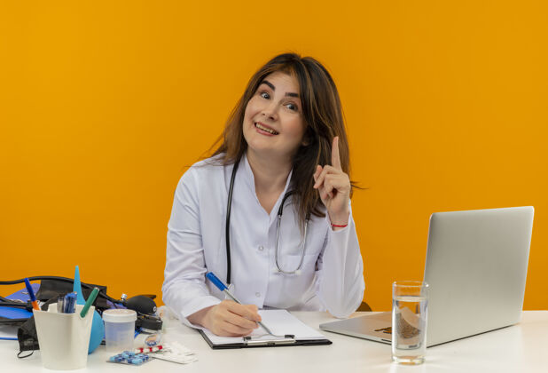 工具微笑的中年女医生穿着医用长袍和听诊器坐在办公桌旁 拿着医疗工具 剪贴板和笔记本电脑拿着钢笔 手指孤立地举起桌子橙色长袍