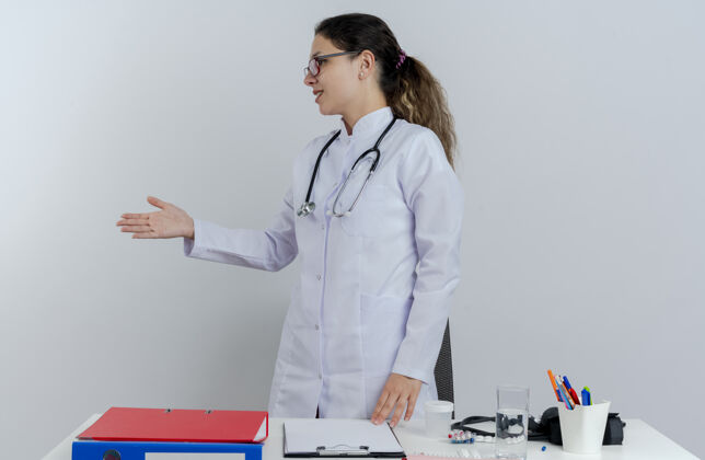 成人年轻的女医生穿着医用长袍 戴着听诊器 戴着眼镜 站在桌子后面 拿着医疗工具 头对侧地打招呼转身长袍医生