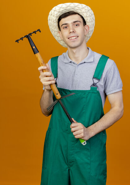 帽子微笑着的年轻男园丁戴着园艺帽拿着耙子锄头耙子在橙色背景上与复制空间隔离持有微笑复制