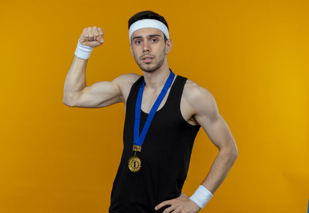 站立戴着头巾 脖子上戴着金牌的年轻运动型男子站在橙色背景下 看着摄像机举起拳头 表情严肃脖子严重年轻