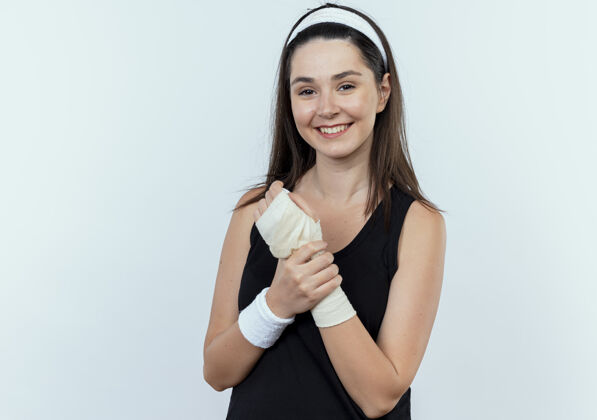 站立戴着头巾的年轻健身女士抚摸着她缠着绷带的手腕 站在白色背景上面带微笑地看着摄像机年轻相机面部