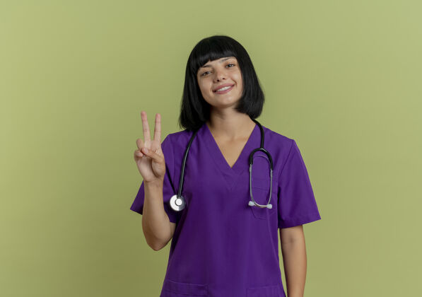 标志微笑的年轻黑发女医生穿着制服 手持听诊器 手势胜利 手势孤立于橄榄绿背景上 留有复印空间胜利复制听诊器
