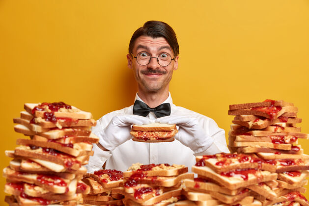 成人快乐友好的侍者有机会品尝美味的三明治 在一堆烤面包的旁边摆姿势 穿着正式的服装 戴着白色的手套 隔离在黄色的墙上吃汉堡的时间到了优雅姿势眼镜
