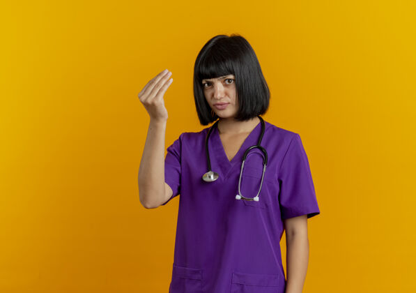 空间笨手笨脚的年轻黑发女医生 穿着制服 用听诊器做手势 把钱的手势孤立在橙色背景上 留有复印空间复制听诊器黑发