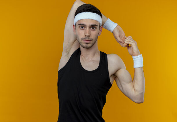 运动戴着头巾的年轻运动型男子站在橙色背景上 看着摄像机伸出手伸展手头带