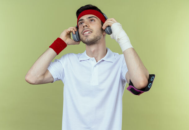 手机戴着头戴式耳机和智能手机臂带的年轻运动型男子 站在绿色背景下 快乐而积极地欣赏着他的音乐积极享受耳机