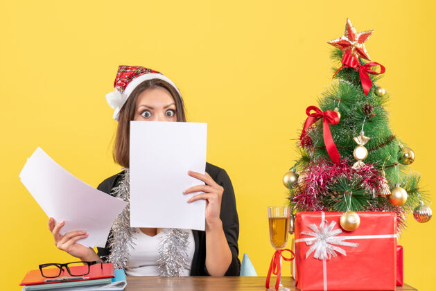 丝带穿着西装 戴着圣诞老人帽 戴着新年装饰品的商务女士惊奇地检查着文件 坐在一张桌子旁 桌子上放着圣诞树圣诞老人支票树