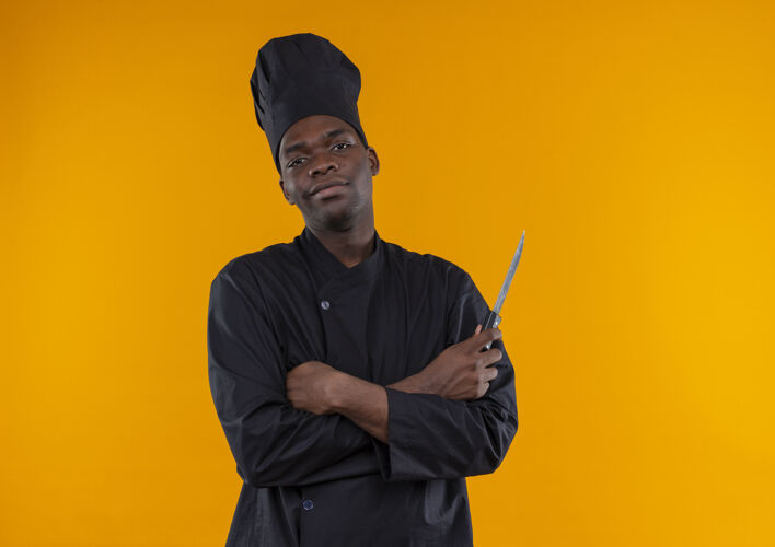 厨师年轻自信的非洲裔美国厨师身着厨师制服 双手交叉持刀 背景为橙色 留有复印空间烹饪刀非裔美国人