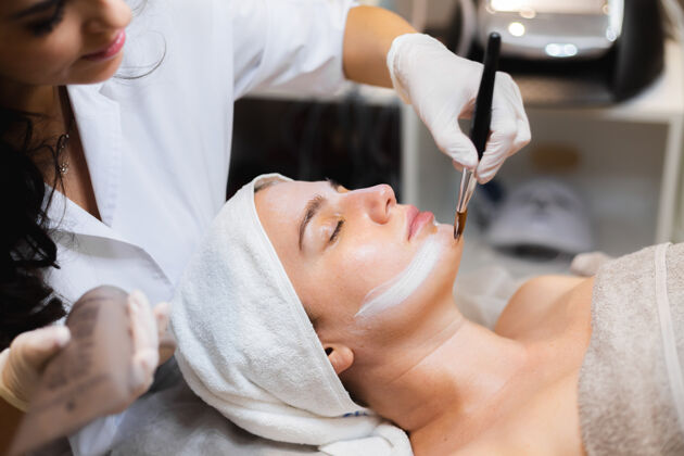 治疗美容师用刷子将白色保湿面膜涂在spa美容院一位年轻女客户的脸上女人健康平静
