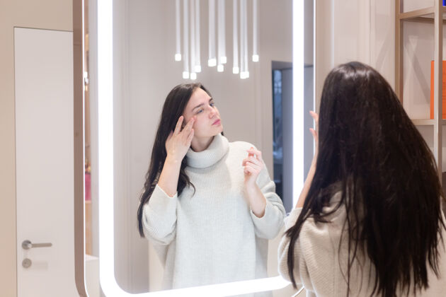 过程在美容院穿毛衣的年轻美女照镜子 抚摸自己的脸 思考即将到来的手术 考虑自己服务皮肤皮肤科