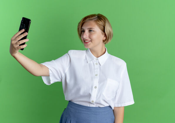 女孩年轻的微笑金发俄罗斯女孩看着手机在绿色的背景与复制空间隔离俄罗斯人电话微笑