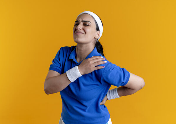 头带年轻疼痛的白人运动妇女戴着头带和腕带举行肩膀和背部橙色背景与复制空间隔离运动疼痛背部