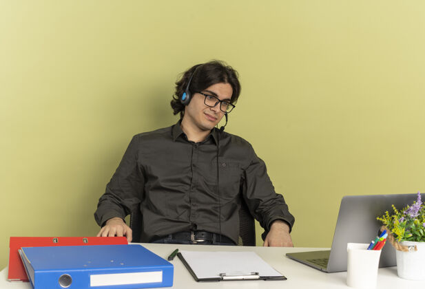 笔记本电脑戴着耳机 戴着眼镜的年轻上班族坐在办公桌旁 使用办公工具 看着隔离在绿色背景上的笔记本电脑 还有复印空间坐着请光学